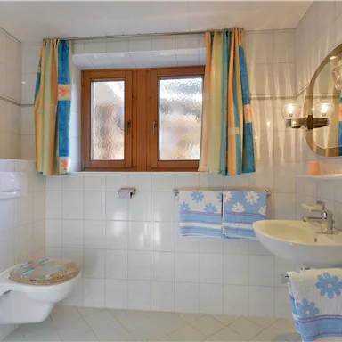 Ein sauberes Badezimmer mit weißen Fliesen und bunten Vorhängen. Es gibt ein Waschbecken, eine Toilette und einen großen Spiegel.