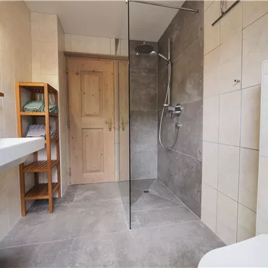 Ein modernes Badezimmer mit einer offenen Dusche und einem schlichten Design. Die Wände sind hell gefliest und es gibt eine Holzablage für Handtücher.