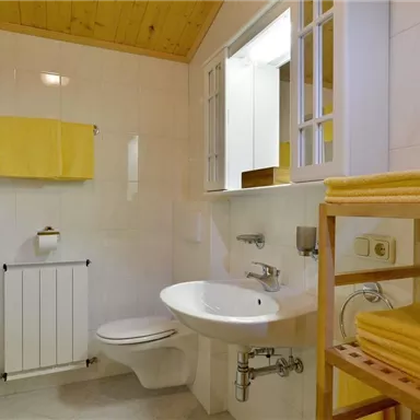 Ein modernes Badezimmer mit weißen Wänden und Holzdecke. Handtücher in gelb hängen zur Dekoration, und es gibt eine Dusche sowie eine Toilette.