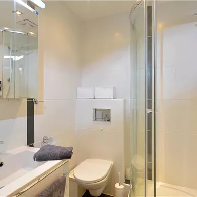 Ein modernes Badezimmer mit Dusche, Waschbecken und Toilette. Die Wände sind hell gefliest und ein großer Spiegel vergrößert den Raum.