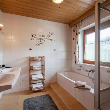Ein helles Badezimmer mit einer Badewanne, Dusche und zwei Waschbecken. An der Wand steht der Schriftzug „Zeit für Entspannung“.