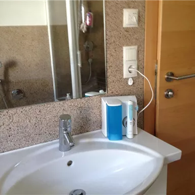 A bathroom with a sink and a mirror. In the background, a shower and a wooden interior door can be seen.