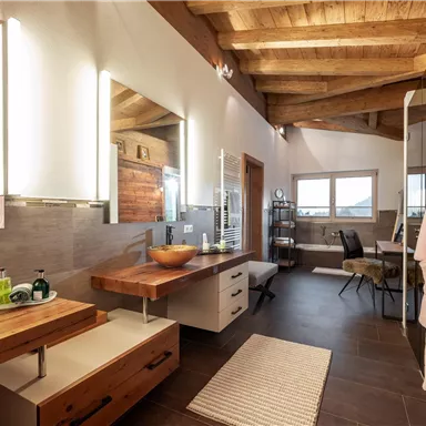 Ein modernes Badezimmer mit Holzdecke und großer Spiegelfläche. Es gibt eine Mischung aus hellen und dunklen Materialien sowie einen Hocker und Pflegeprodukte auf der Holzkommode.