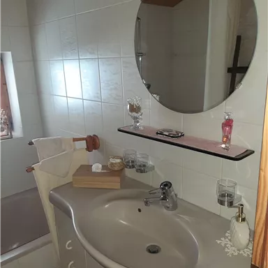 Ein kleines Badezimmer mit einem runden Spiegel über dem Waschbecken. Auf dem Regal befinden sich Pflegeprodukte und ein Handtuchhalter.