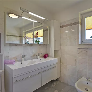 Ein modernes Badezimmer mit hellem, elegantem Design. Es gibt einen Doppelwaschtisch, einen großen Spiegel und eine Pflanze auf der Fensterbank.