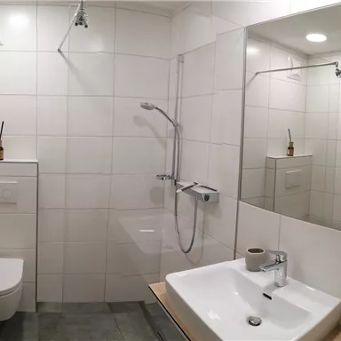 Ein modernes Badezimmer mit einer Dusche, einem Waschbecken und einer Toilette. Die Wände sind mit weißen Fliesen verkleidet und es gibt einen großen Spiegel.