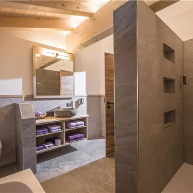 A modern bathroom with a shower and a bathtub. The walls are covered with gray tiles and there is a large mirror area.