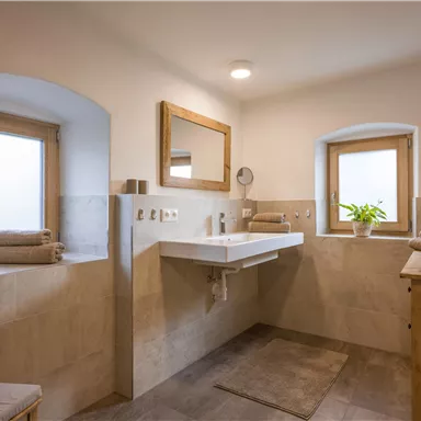 Ein modernes Badezimmer mit hellen Wänden und einem großen Waschbecken. Das Zimmer hat Fenster, durch die viel Licht kommt, und Dekoration aus Holz.