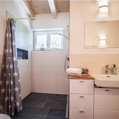 A modern bathroom with a shower and a sink. The walls are light tiled and there is a large mirror.