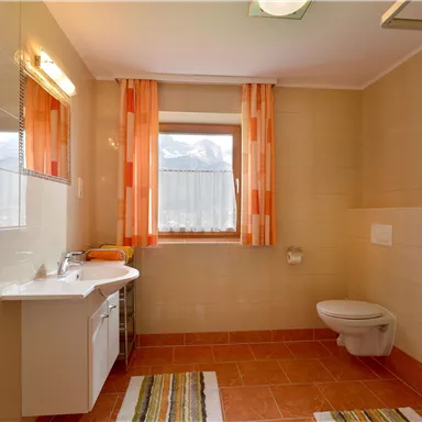 A modern bathroom with orange curtains and a large window. There is a shower, a toilet, and a sink unit.
