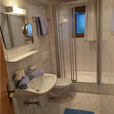 Ein modernes Badezimmer mit Dusche, Waschbecken und WC. Die Wände sind gefliest, und es gibt einige Handtücher und Pflegeartikel auf den Regalen.