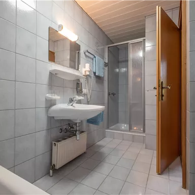 Ein modernes Badezimmer mit grauen Fliesen. Es gibt eine Dusche, ein Waschbecken und einen Spiegel sowie Handtücher auf einem Regal.