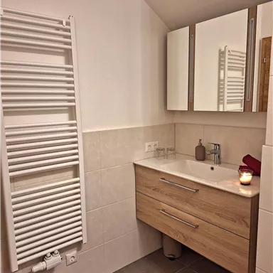 Ein modernes Badezimmer mit einem Holzwaschbecken, Spiegel und Handtuchheizkörper. Eine rote Badematte und eine Kerze sorgen für eine gemütliche Atmosphäre.