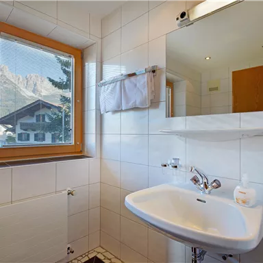 Ein modernes Badezimmer mit einem Waschbecken, einem Spiegel und einem Fenster, das einen Blick auf die Berge bietet. Die Wände sind hell gefliest und der Raum wirkt hell und einladend.