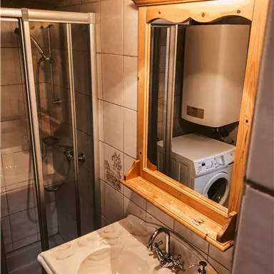 Ein kleines Badezimmer mit einer Duschkabine, einem Waschbecken und einem Spiegel. Im Hintergrund steht eine Waschmaschine.