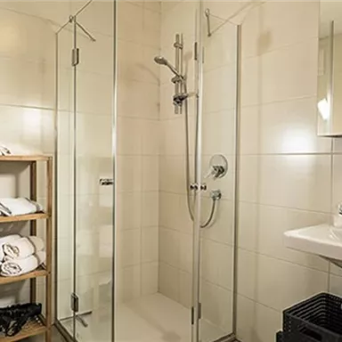 Ein modernes Badezimmer mit einer Dusche aus Glas und einem Waschbecken. Handtücher sind auf einem Holzregal ordentlich gestapelt.