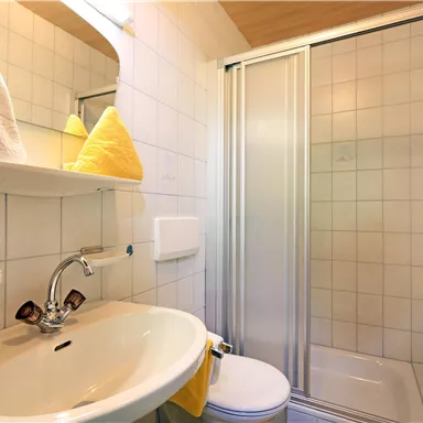 Ein sauberes Badezimmer mit einem Waschbecken, einer Dusche und weißen Fliesen. Gelbe Handtücher und Toilettenartikel verleihen dem Raum eine einladende Atmosphäre.