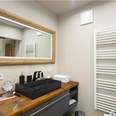 A modern bathroom with a large mirror and a wooden table. A towel radiator hangs on the wall and next to the sink are care products.