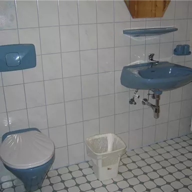 A bathroom with blue sanitary installations and white tiles. On the left is the toilet and on the right is a sink with a small shelf above it.