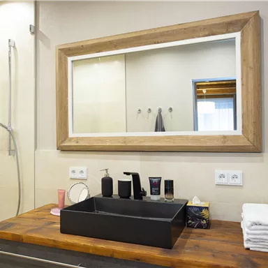 A modern bathroom with a large mirror surface and an elegant sink made of black material. The wooden surface and the shower cabin give the room an inviting atmosphere.