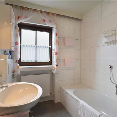 Ein modernes Badezimmer mit einer Badewanne und einem Waschbecken. Es gibt ein Fenster mit Vorhängen und helle, geflieste Wände.