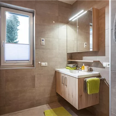 A modern bathroom with a wood vanity and a large mirror. Light gray tiles and green towels create a fresh atmosphere.