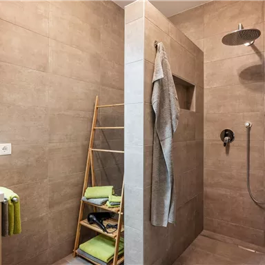 Modern shower with gray tiles and a rectangular shower area. A towel rack and toiletries are visible in the room.