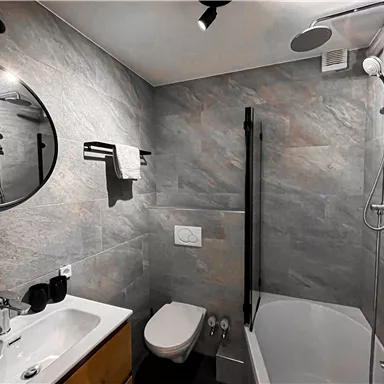 Ein modernes Badezimmer mit grau gefliesten Wänden. Es verfügt über eine Badewanne, ein Waschbecken und eine Dusche sowie einen großen Spiegel.