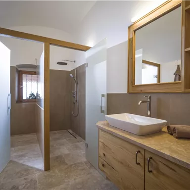 A modern bathroom with a shower and a ceramic sink. The furnishings are made of wood and the walls are light-colored.