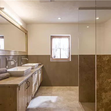 A modern bathroom with wooden cabinets and two sinks. The shower is separated by glass, and there is plenty of natural light from a window.