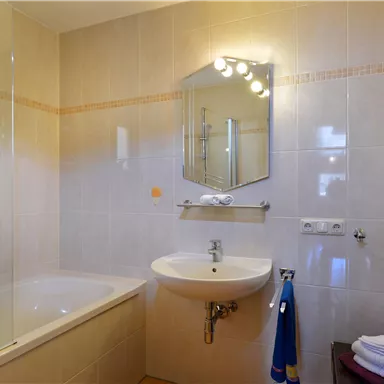A modern bathroom with a bathtub and a sink. The walls are tiled in light colors and there are fresh towels as well as a small plant.
