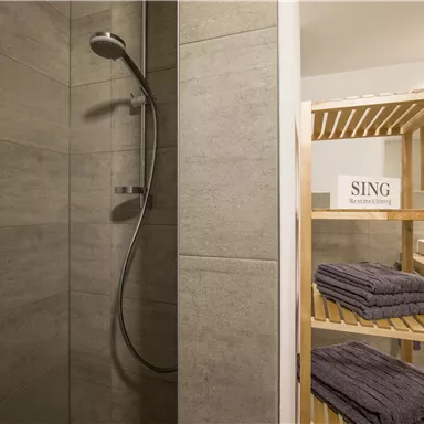 Ein modernes Badezimmer mit einer Dusche und einem Holzregal. Auf dem Regal liegen Handtücher und ein Schild mit der Aufschrift „SING“.