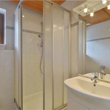 Ein modernes Badezimmer mit einer Duschkabine und einem Waschbecken. Die Wände sind mit weißen Fliesen gestaltet und es gibt viel Tageslicht.