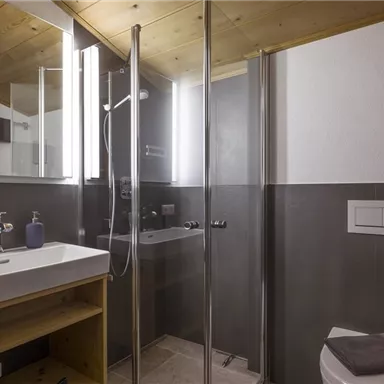 Ein modernes Badezimmer mit einer Glasdusche und einer schlichten Waschtischanlage. Die Wände sind in dunklem Grau gehalten, während die Decke aus hellem Holz besteht.