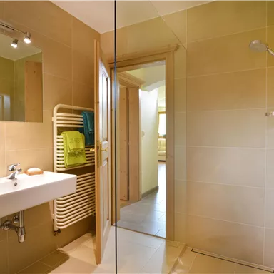 Ein modernes Badezimmer mit einer Dusche und einem Waschbecken. Die Wände sind in warmen Farbtönen gehalten, und Handtücher sind an einem Heizkörper aufgehängt.