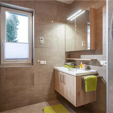 A modern bathroom with a wood vanity and a large mirror. Light gray tiles and green towels create a fresh atmosphere.