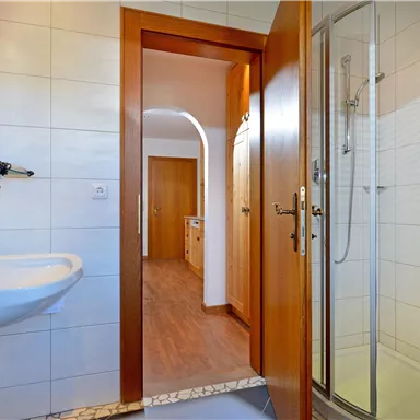 Ein modernes Badezimmer mit einem Waschbecken und einer Duschkabine. Die Wände sind gefliest und es gibt einen Holzrahmen an der Tür.