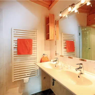 Ein modernes Badezimmer mit einem Doppelwaschtisch und einem großen Spiegel. An der Wand hängen weiße Handtücher und es gibt einen Duschbereich mit Glasabtrennung.