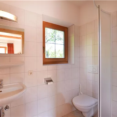 Ein helles Badezimmer mit weißen Fliesen und einem Fenster, das Tageslicht hereinlässt. Es gibt ein Waschbecken, eine Dusche und eine Toilette.