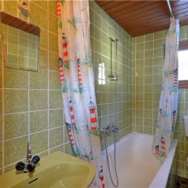 Ein einfaches Badezimmer mit grünen Fliesen. Es gibt eine Badewanne, eine Toilette und einen kleinen Spiegel.