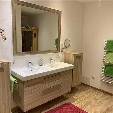 A modern bathroom with a double sink and a large mirror. Wooden shelves and towels in shades of green and red give the room a fresh atmosphere.