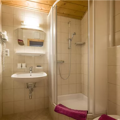 Ein modernes Badezimmer mit einer Dusche und einem Waschbecken. Die Wände sind hell gefliest und Handtücher in lila Farbe sind vorhanden.