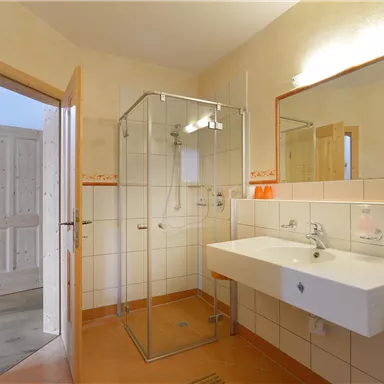 Ein modernes Badezimmer mit Dusche, Waschbecken und Spiegel. Die Wände sind hell gefliest und es gibt eine offene Tür zu einem weiteren Raum.