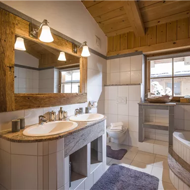 Ein modernes Badezimmer mit zwei Waschbecken und einem großen Spiegel. Der Raum hat Holzelemente und ein Fenster, das viel Licht hereinlässt.