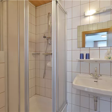 Ein einfaches Badezimmer mit einer Dusche, einem Waschbecken und einem Spiegel. Die Wände sind gefliest und es gibt grundlegende Toilettenartikel.