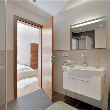 Ein modernes Badezimmer mit Dusche, Waschbecken und großer Spiegelleuchte. Die Tür öffnet sich zu einem angrenzenden Schlafzimmer.