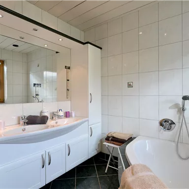 Ein modernes Badezimmer mit einer Badewanne und einem großen Spiegel. Die Wände sind gefliest und die Ausstattung ist in Weiß gehalten.