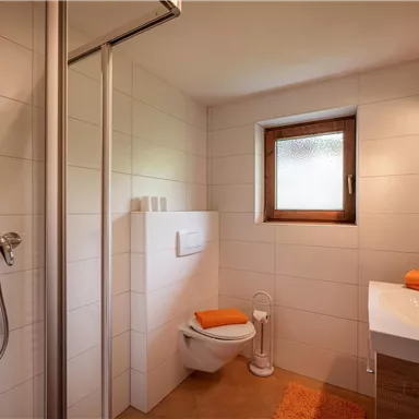 Ein modernes Badezimmer mit Dusche, Toilette und Waschbecken. Helle Fliesen und orangefarbene Handtücher schaffen eine angenehme Atmosphäre.