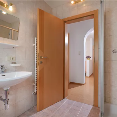 A bright bathroom with a sink, a mirror, and a shower. The doors are made of wood and lead to another room.