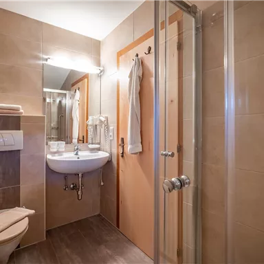 Ein modernes Badezimmer mit einer Dusche, einem Waschbecken und einem WC. Die Wände sind hell gefliest und eine große Spiegelhatte ist vorhanden.